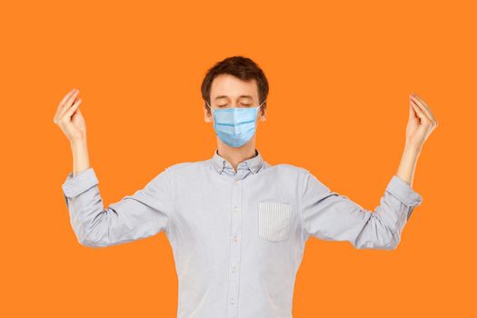 Yoga and meditation. Portrait of calm young worker man with surgical medical mask standing with closed eyes and meditating. indoor studio shot isolated on orange background.