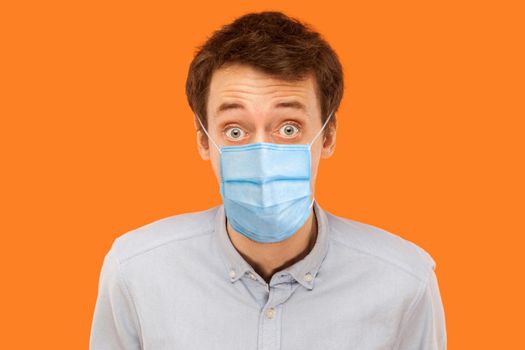 Closeup portrait of shocked young worker man with surgical medical mask standing and looking at camera with big eyes. indoor studio shot isolated on orange background.