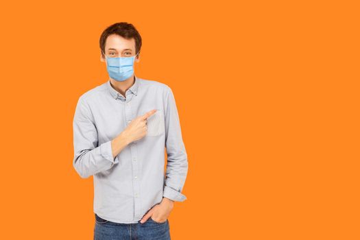 Portrait of happy young worker man with surgical medical mask standing, pointing and showing background empty copy space and smiling. indoor studio shot isolated on orange background.