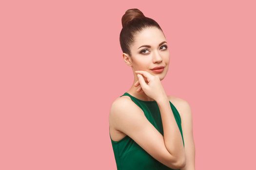 Closeup portrait of happy beautiful young woman with bun hairstyle and makeup in green dress standing touching her face and looking away with smile. indoor studio shot, isolated on pink background.
