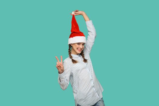 New year concept. happy funny beautiful girl in striped light blue shirt standing and holding red christmas hat, showing victory or peace sign looking at camera. indoor isolated on green background.