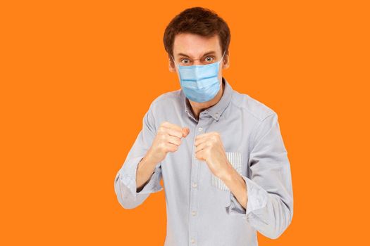 Portrait of angry young worker man with surgical medical mask standing in boxing fists and looking at camera and ready to attack against virus. indoor studio shot isolated on orange background.