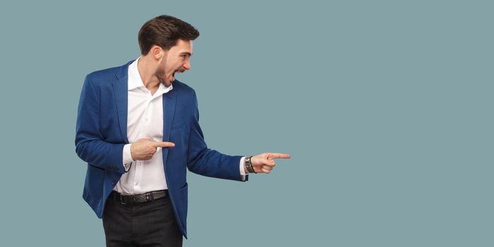 Handsome amazed bearded man in blue suit standing and pointing at background and screaming and looking at copy space. studio, indoor shot isolated on blue background.
