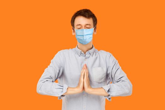 Yoga and meditation. Portrait of calm young worker man with surgical medical mask standing with closed eyes and palm hands meditating. indoor studio shot isolated on orange background.