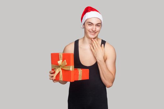 Cunning man in red hat holding gift box touching his chin and toothy smiling looking at camera with funny face. indoor, studio shot, isolated on gray background