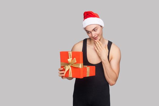 New year concept. Man in red hat holding red gift box and looking inside with amazed funny face. Indoor, studio shot, isolated on gray background