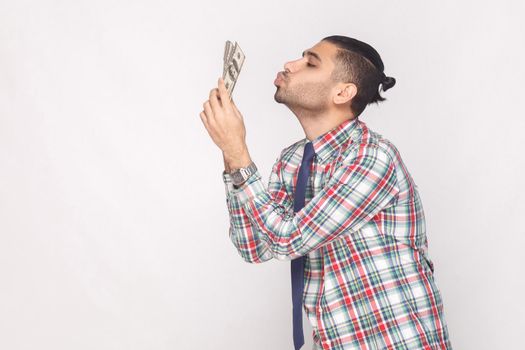 Profile side view of cheerful rich handsome bearded young stylish businessman in colorful checkered shirt standing and air kissing fan of dollars. Indoor, studio shot, isolated on grey background.