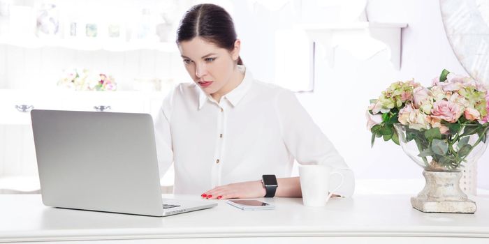 Young business woman wearing smartwatch using laptop computer. beautiful assistant with dark brown hair, blue eyes and white shirt working on laptop in the light white office. looking at display and working.