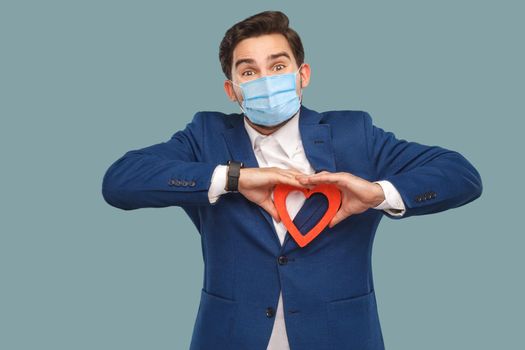 Handsome man with surgical medical mask in blue jacket standing and holding red heart shape and looking at camera. medicine and health care concept. Indoor, studio shot isolated on blue background