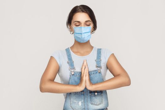 relaxation and yoga meditation. Portrait of calm young brunette woman with surgical medical mask in denim overalls standing in yoga pose and meditating. indoor studio shot isolated on gray background.