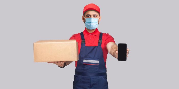 Delivery on quarantine. This is For you! Young man with surgical medical mask in blue uniform and red t-shirt standing, holding cardboard box and showing smart phone display on grey background.