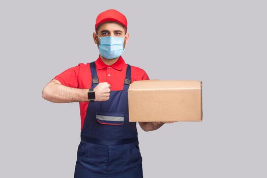 Delivery on quarantine. Ontime service! man with surgical medical mask in blue uniform and red t-shirt standing, holding delivery box and showing watch on grey background. Indoor shot, isolated,