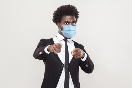 Portrait of serious or shocked young handsome worker man wearing black suit with surgical medical mask standing looking and pointing and camera. indoor studio shot isolated on gray background.