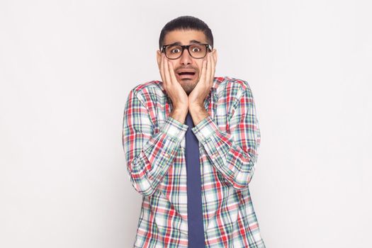 Shocked bearded businessman in colorful checkered shirt, blue tie and black eyeglasses standing, touching face and looking at camera with scared face. indoor studio shot, isolated on grey background.