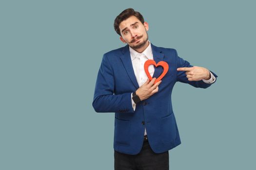 Sad unhappy broken heart man in blue jacket and white shirt, standing and holding red heart shape and looking at camera and pointing finger. Indoor, studio shot isolated on light blue background