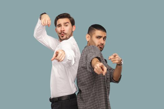 Now is your time to win. portrait of two handsome bearded friends or partners standing, looking and pointing at camera with funny amazed faces. indoor studio shot, isolated on light blue background.