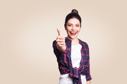 Young happy girl with casual style and bun hair thumbs up her finger, on beige blank wall with copy space looking at camera with toothy smile. focus on face.