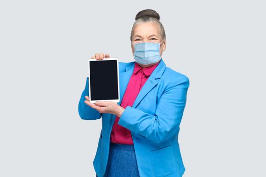 Aged woman with surgical medical mask holding and showing tablet screen. Grandmother in light blue suit with collected gray hair bun hairstyle. indoor studio shot, isolated on gray background