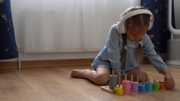Happy Little Preschool Toothless Girl Playing With Colored Wooden Toy. Kids Learn To Count By Playing Teaches Numbers At Home. Child Listening To Music In Big White Headphones. Childhood, Education.