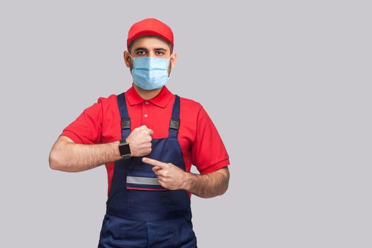 We do ontime. Young man with surgical medical mask in blue overall and red t-shirt standing and showing time on his wrist watch with smile. Grey background, indoor, studio shot, isolated