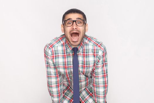 Happy surprised bearded businessman in colorful checkered shirt, blue tie and black eyeglasses standing and looking at camera with amazed face and open mouth. studio shot, isolated on grey background