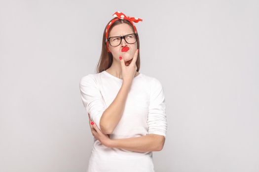 portrait of beautiful emotional young woman in white t-shirt with freckles, black glasses, red lips and head band touching her chin and thinking. indoor studio shot, isolated on light gray background.
