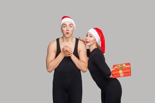 Couple in christmas hat. Shocked man looking at camera. woman holding and hiding red gift box and want to make him surprise. Indoor, studio shot, isolated on gray background