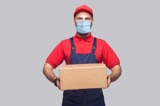 Delivery on quarantine. Portrait of young man with surgical medical mask in blue uniform and red t-shirt standing and holding the cardboard box on grey background. Indoor, studio shot, isolated,