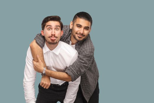 portrait of two handsome happy bearded friends standing, enjoying, hugging and looking at camera with smile and funny face and tongue out. indoor studio shot, isolated on light blue background.