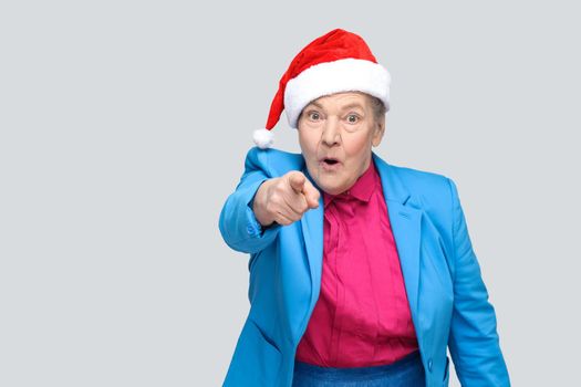 Amazed grandmother in colorful casual style, blue suit and christmas red cap standing and looking and pointing finger at camera with shocked face. indoor, studio shot, isolated on gray background