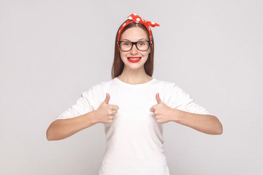 I am satisfied, thumbs up. portrait of beautiful emotional young woman in white t-shirt with freckles, black glasses, red lips and head band. indoor studio shot, isolated on light gray background.