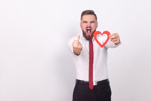 Fuck you, love! Angry man showing hate sign, holding heart. Indoor, studio shot, isolated on gray background