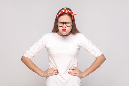 angry thinkful, raised hands. portrait of beautiful emotional young woman in white t-shirt with freckles, black glasses, red lips and head band. indoor studio shot, isolated on light gray background.