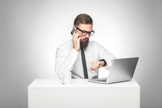 Thoughful bearded young worker in white shirt and black tie are sitting in office on desk and talking with partner on phone also checking time on his own hand watch, planning meeting. Gray background