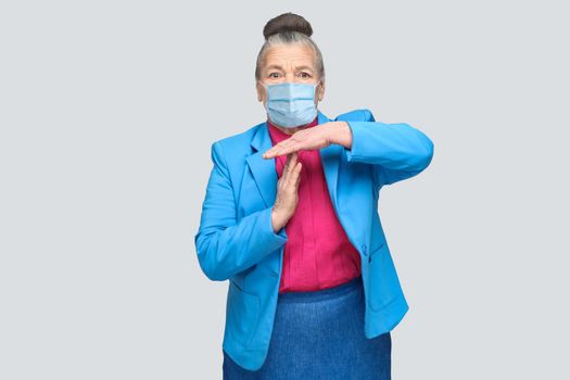 Aged woman with surgical medical mask showing time out sign. Grandmother with light blue suit and pink shirt standing with collected bun gray hair. indoor studio shot, isolated on gray background