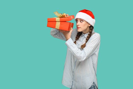 New year concept. cunning beautiful girl in striped light blue shirt in red christmas cap standing holding red gift box, unboxing and looking inside. indoor, studio shot isolated on green background.