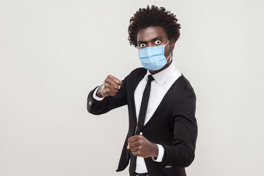 Portrait of angry man wearing black suit with surgical medical mask standing with boxing fists and ready to attack or defence. indoor studio shot isolated on gray background.