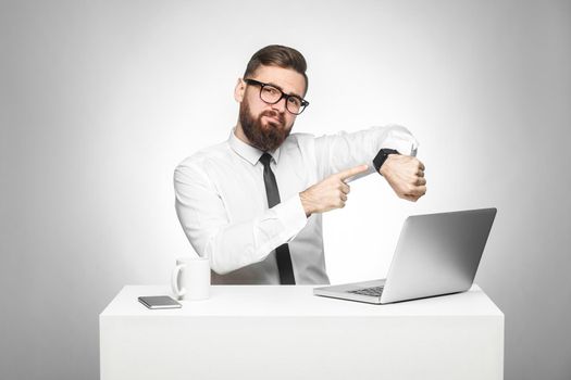 Portrait of warning handsome bearded young boss in white shirt and black tie are sitting in office and pointing finger to him hand watch showing you time has come. Indoor, studio shot, gray background