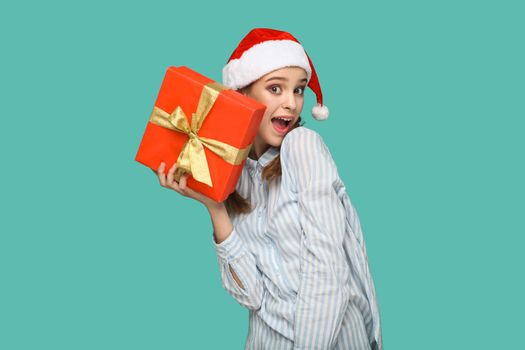 New year concept. wondered beautiful girl in striped light blue shirt in red christmas cap standing, holding red gift box and looking at camera with amazed face. indoor isolated on green background.