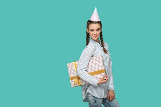Happy funny girl in striped blue shirt standing with dotted gift box and birthday cap on her head, looking at camera with safisfied smiley face, Indoor studio shot, isolated on green background