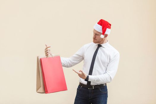 Black friday in winter. Happiness red head businessman in santa hat , holding shopping bags on hands, pointing fingers and looking at gifts. Indoor shot, light orange background