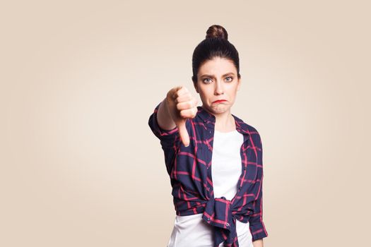 Dislike. Young unhappy upset girl with casual style and bun hair thumbs down her finger, on beige blank wall with copy space looking at camera with toothy smile. focus on face.