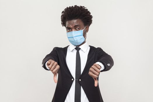 Dislike. Portrait of dissatisfied young worker man wearing black suit with surgical medical mask standing holding thumbs down and looking ar camera. indoor studio shot isolated on gray background.