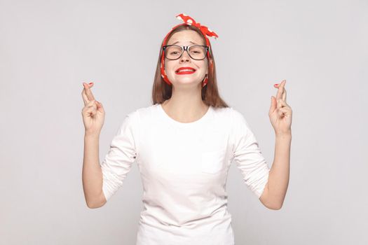 hopeful portrait of beautiful emotional young woman in white t-shirt with glasses, red lips and head band looking at camera with crossed finger. indoor shot, isolated on light gray background.