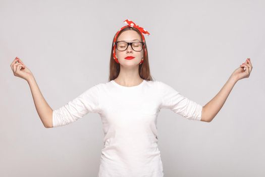 yoga relaxed raised hands portrait of beautiful emotional young woman in white t-shirt with freckles, black glasses, red lips and head band. indoor studio shot, isolated on light gray background.