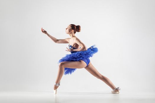 Young beautiful ballerina with bun collected hair wearing blue dress and pointe shoes dancing gracefully isolated on white background. indoor, studio shot.