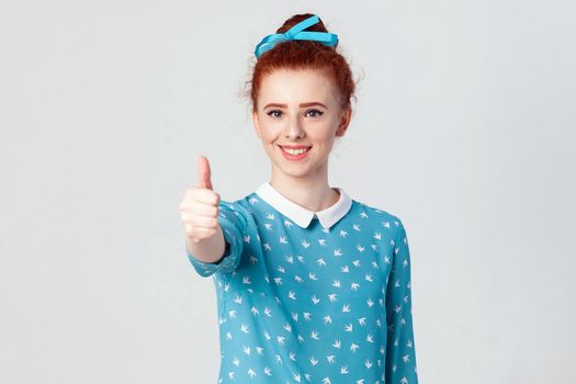 Young happy cheerful redhead girl showing thumb up and toothy smile. Isolated studio shot on gray background