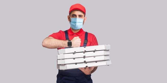 In time delivery on quarantine! man with surgical medical mask in uniform and red t-shirt standing, holding stack of cardboard pizza boxes on grey background. Indoor, studio shot, isolated, copy space