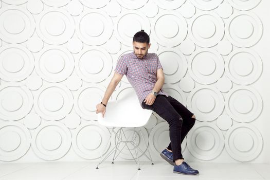Young bearded fashion model in casual style is posing near white circle wall background. studio shot. sitting on chair and looking down.