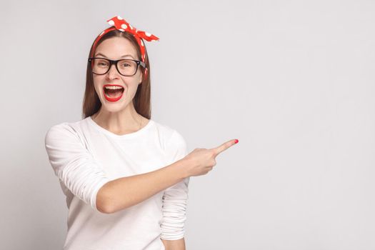 happiness, wondering portrait of beautiful emotional young woman in white t-shirt with freckles, glasses, red lips and head band pointing copyspace. indoor shot, isolated on light gray background.